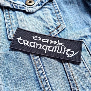 May include: A rectangular black patch with the words "Dark Tranquillity" embroidered in white script. The patch is placed on a light blue denim jacket, with visible stitching and a metal button.