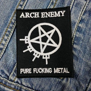 May include: Black square patch with white text and a pentagram design. The text at the top reads "ARCH ENEMY", and the text at the bottom reads "PURE FUCKING METAL". The patch is sewn onto a denim jacket.