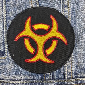 May include: A black and yellow biohazard symbol patch with red stitching. The symbol is a stylized triangle with three circles.