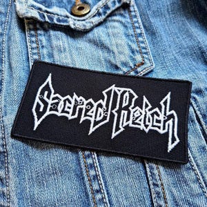 Op de afbeelding: Zwarte en witte geborduurde patch met de tekst "Sacred Reich" op een spijkerjack.