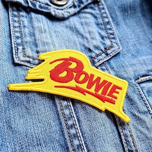 May include: A yellow embroidered patch with the name "Bowie" in red script. The patch is in a stylized shape and is affixed to a denim jacket. The patch is a vibrant accessory.