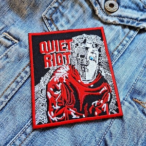 Könnte beinhalten: Rechteckiger Aufnäher mit den Worten "QUIET RIOT" in roten Blockbuchstaben. Der Aufnäher zeigt eine rot-weiße Illustration eines Gesichts auf schwarzem Hintergrund mit rotem Rand. Der Aufnäher befindet sich auf einer Jeansjacke.