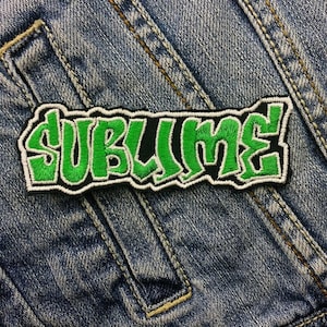 Op de afbeelding: Groen en zwart geborduurd patch met het woord "Sublime" in een gestileerd lettertype. Het patch is genaaid op een blauw denim jack.