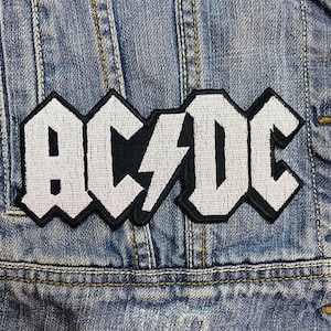 Może przedstawiać: Haftowana naszywka z nazwą zespołu "AC/DC" w białych literach blokowych, czarną błyskawicą i obramowaniem. Naszywka jest przyszyta do niebieskiej kurtki dżinsowej, prezentując klasyczną estetykę rock and rolla.