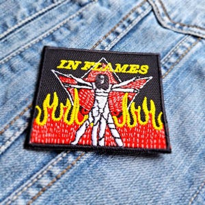 Op de afbeelding: Een zwarte, vierkante patch met de tekst "IN FLAMES" in het geel. De patch toont een gestileerde figuur met uitgestrekte armen, rode vleugels en gele vlammen aan de onderkant. De patch is geplaatst op een denim achtergrond.