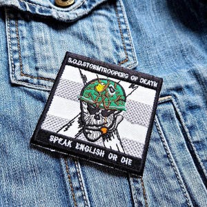 Pode incluir: Um remendo preto e branco com uma caveira usando um capacete militar verde com um pássaro amarelo no topo. O texto "S.O.D. STORM TROOPER OF DEATH" está acima da caveira e "SPEAK ENGLISH OR DIE" está abaixo da caveira.