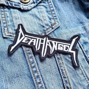 Può includere: Una toppa ricamata in bianco e nero con il testo "Death Angel" su una giacca di jeans blu.