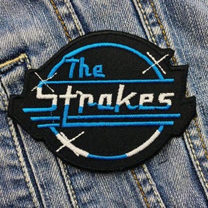 May include: A black and blue embroidered patch with the text "The Strokes" in a white circle with a blue outline.