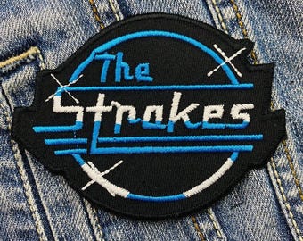May include: A black and blue embroidered patch with the text "The Strokes" in a white circle with a blue outline.