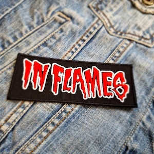 May include: A black and red embroidered patch with the text "IN FLAMES" in a dripping font style. The patch is sewn onto a blue denim jacket.