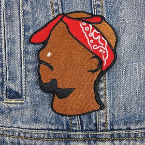 Puede incluir: Parche bordado que representa el perfil de una persona con piel morena, un bigote negro y un pañuelo rojo con diseños de cachemira blancos. El parche está cosido sobre una chaqueta vaquera.