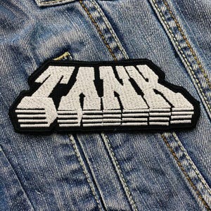 Könnte beinhalten: Weißer gestickter Patch mit dem Wort "TANK" in fetten Buchstaben auf schwarzem Hintergrund. Der Patch hat ein gestreiftes Design unter dem Wort.