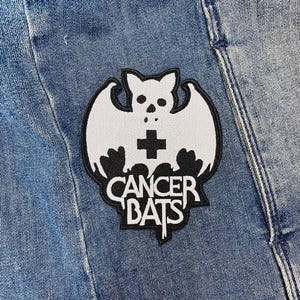 以下が含まれることがあります： 青いデニムジャケットに、胸に十字架、黒い文字で「Cancer Bats」と書かれた白いコウモリが描かれている。