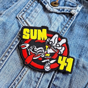 Puede incluir: Un parche bordado negro y rojo con las palabras "SUM 41" en amarillo. El parche presenta un personaje de dibujos animados con una camisa a rayas rojas y blancas y una mano negra y blanca.