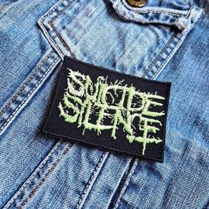 Könnte beinhalten: Ein schwarzer Patch mit den Worten "Suicide Silence", die in grünem Garn gestickt sind.