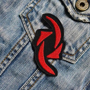 Puede incluir: Parche bordado rojo y negro con un diseño estilizado. El parche está cosido en una chaqueta vaquera azul.