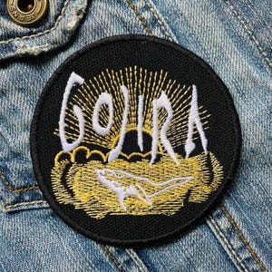 Peut inclure: Patch brodé noir avec le mot "GOJIRA" en lettres blanches, entouré d'un motif de soleil jaune et d'une silhouette blanche de requin.