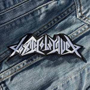 Peut inclure: Patch brodé blanc avec le mot "SLAUGHTER" dans une police stylisée sur un fond noir. Le patch est cousu sur une veste en jean bleue.