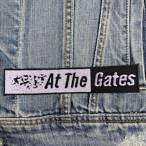Puede incluir: Un parche bordado en blanco y negro con el texto "At The Gates" sobre un fondo morado claro. El parche está cosido en una chaqueta vaquera azul.