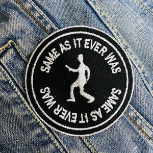 May include: Black and white embroidered patch with the text "Same as it ever was" and a stick figure walking. The patch is sewn onto a blue denim jacket.