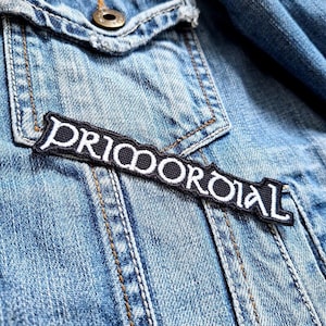 Pode incluir: Um close-up de uma jaqueta jeans com um patch bordado em preto e branco. O patch diz "PRIMORDIAL" em letras brancas estilizadas. A jaqueta é azul clara com costuras visíveis e um botão de metal.