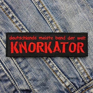 Puede incluir: Parche rectangular negro con texto rojo sobre fondo vaquero azul. El parche dice "deutschlands meiste band der welt" encima de la palabra "KNORKATOR". El parche es probablemente un accesorio de ropa.