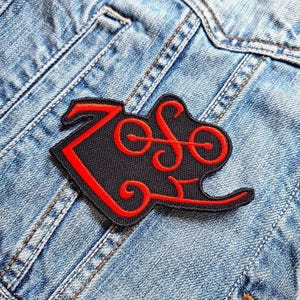 May include: A black and red embroidered patch with the word "Zoso" in a stylized font. The patch is sewn onto a blue denim jacket.