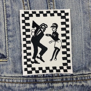 Peut inclure: Patch brodé noir et blanc de deux personnages dansant sur un fond à carreaux. Le patch est cousu sur une veste en jean bleue.
