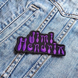 May include: A black patch embroidered with the name "Jimi Hendrix" in purple script. The patch is set against a denim jacket, showing stitching and a pocket. The patch has a black border.
