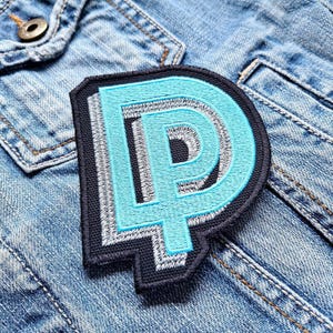 Op de afbeelding: Een blauwe en witte geborduurde patch met de letter "D" in een vetgedrukt, gestileerd lettertype. De patch is genaaid op een spijkerjack.