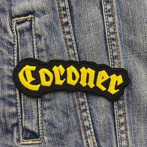 Peut inclure: Un écusson brodé noir et jaune avec le mot "Coroner" en police gothique.