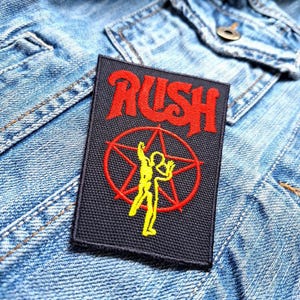 May include: A black fabric patch with the word "RUSH" in red lettering. A red star with a yellow silhouette of two figures is in the centre of the patch.