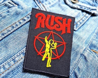 Peut inclure: Un patch en tissu noir avec le mot "RUSH" en lettres rouges. Une étoile rouge avec une silhouette jaune de deux personnages est au centre du patch.