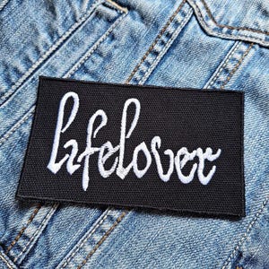 May include: Black fabric patch with white embroidered text that reads "Lifelover".