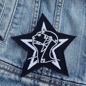 Könnte beinhalten: Schwarz-weiß gestickter Patch mit zwei Profilgesichtern in einem fünfzackigen Stern. Der Patch ist auf eine blaue Jeansjacke genäht.