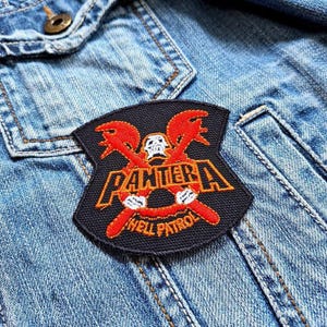 Op de afbeelding: Zwarte en oranje Pantera "Hell Patrol" patch op een spijkerjasje. De patch toont een schedel, gekruiste bijlen en de naam van de band in oranje letters. De patch is rechthoekig met een gebogen bovenkant.