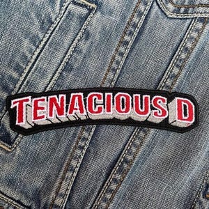 Könnte beinhalten: Ein gestickter schwarzer und weißer Aufnäher mit den Worten "Tenacious D" in roten Buchstaben. Der Aufnäher hat einen 3D-Effekt und ist auf eine Jeansjacke genäht.