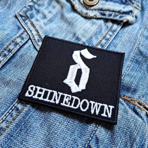 Può includere: Toppa ricamata in bianco e nero con il nome della band "SHINEDOWN" e una lettera "S" stilizzata in bianco.
