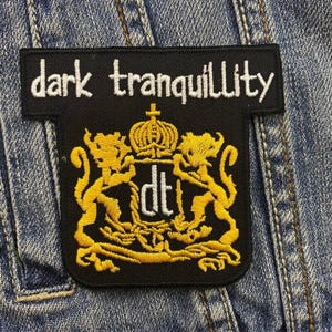 May include: Black embroidered patch with the text "dark tranquility" and a gold crest with the letters "dt" in white.