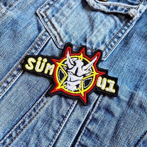 May include: A black, red, and yellow embroidered patch with a white devil's head inside a star. The patch says "SÜM" and "UI" on either side of the star.
