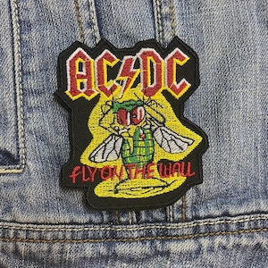 Op de afbeelding: Geborduurde patch met het AC/DC-logo in rood en geel, en een cartoon vlieg illustratie. De tekst "FLY ON THE WALL" staat eronder. De patch is op een spijkerjasje.