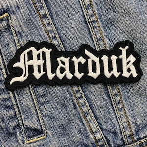 Puede incluir: Un parche bordado en blanco y negro con la palabra "Marduk" en fuente gótica. El parche es rectangular con un borde curvo y está colocado sobre una chaqueta vaquera.