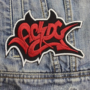 May include: Embroidered patch with the band name "AC/DC" in red, stylized lettering on a black background. The patch has a white border and is set against a denim jacket.