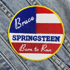 Könnte beinhalten: Ein runder, gestickter Aufnäher mit einem rot-weiß-blauen amerikanischen Flaggenmuster. Der Text "Bruce Springsteen Born to Run" ist in Weiß auf den Aufnäher gestickt.