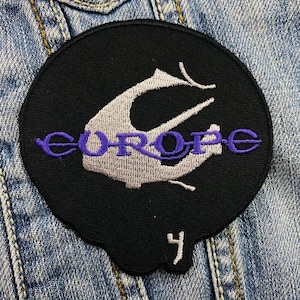 Op de afbeelding: Een zwarte en paarse geborduurde patch met het woord "EUROPE" en een gestileerd vogelontwerp. De patch heeft een nummer "4" in de rechterbenedenhoek.