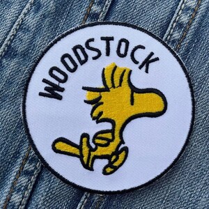 Op de afbeelding: Witte ronde patch met zwarte tekst die "WOODSTOCK" zegt en een gele cartoonvogel met zwarte contouren.