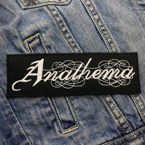 May include: Black embroidered patch with white cursive lettering that reads "Anathema" on a denim background.