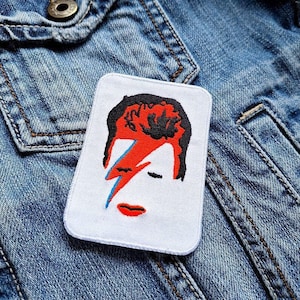 Può includere: Toppa ricamata con un ritratto stilizzato con capelli rossi, un fulmine blu e rosso e labbra rosse. La toppa è rettangolare con angoli arrotondati ed è posta su una giacca di jeans.