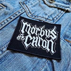 May include: A black, square patch featuring the white embroidered words "Morbus Charon". The patch is displayed on a blue denim jacket. The text is in a gothic font.