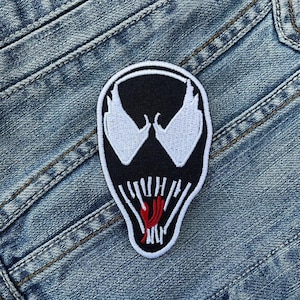 May include: Black and white embroidered patch of Venom's face with a red tongue. The patch is on a blue denim background.